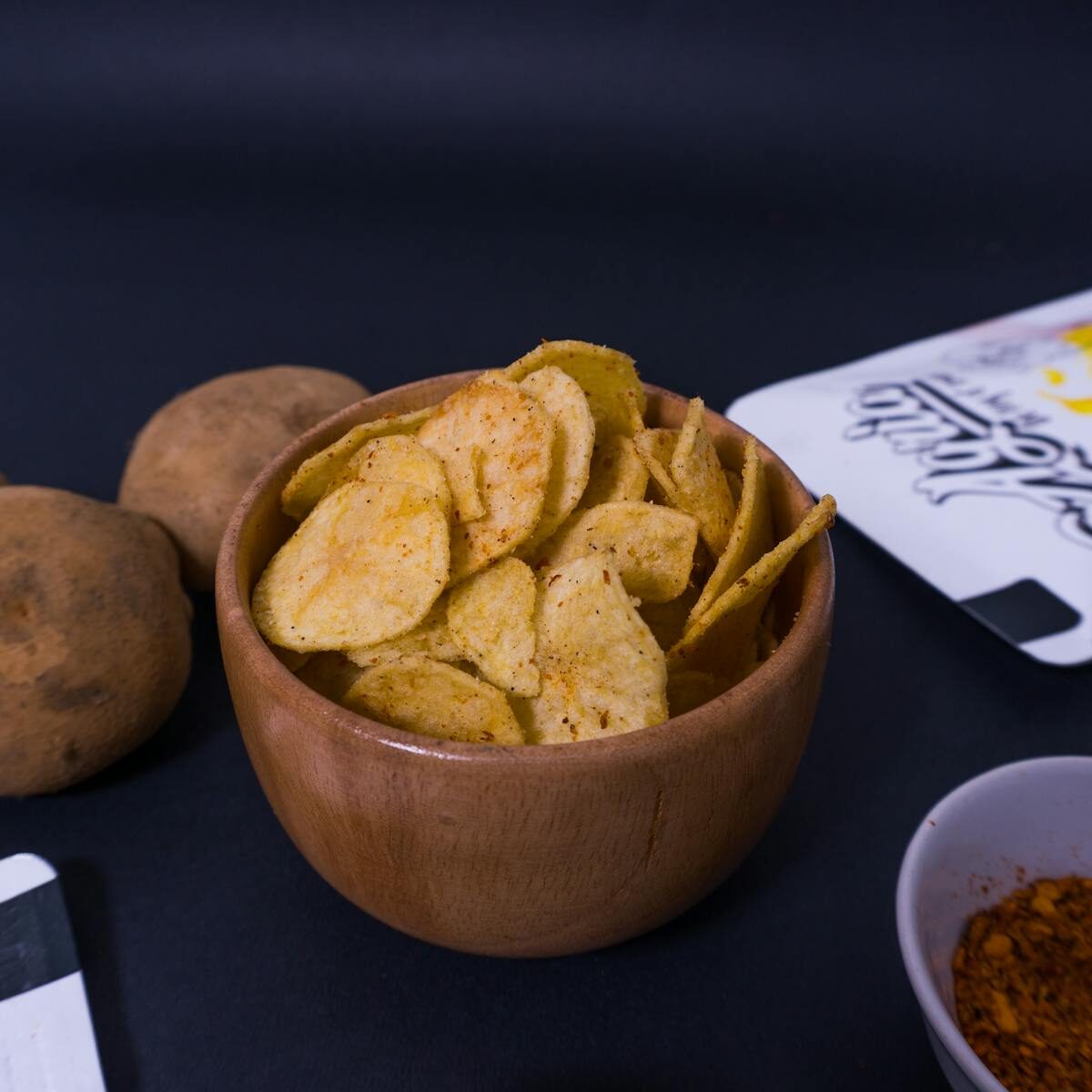 Crispy potato chips in a rustic wooden bowl with seasoning and potatoes nearby.