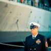 A US Navy officer in dress uniform standing on a ship's deck, representing military professionalism.