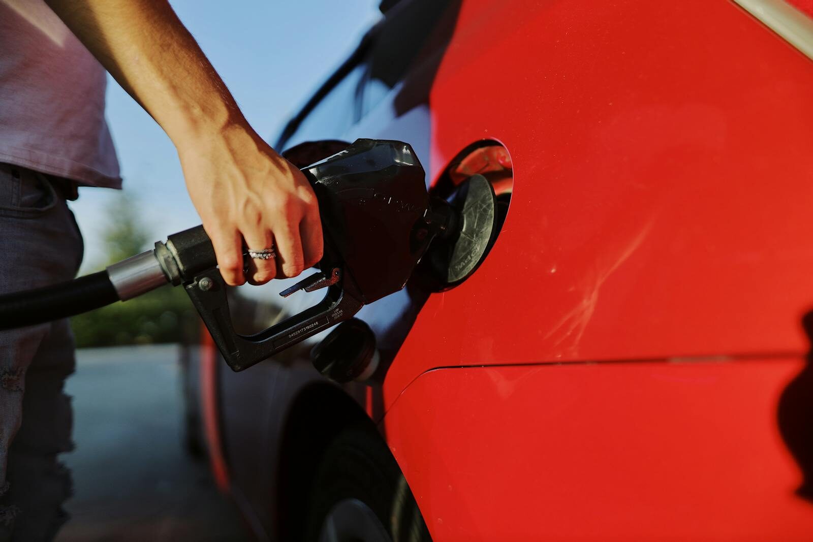 Close-up of a person refueling a red car at an outdoor gas station during the day.