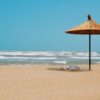Peaceful sandy beach scene with straw umbrella and empty loungers under a clear blue sky.