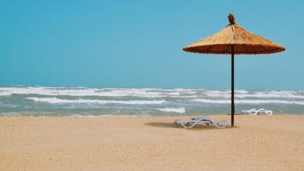 Peaceful sandy beach scene with straw umbrella and empty loungers under a clear blue sky.