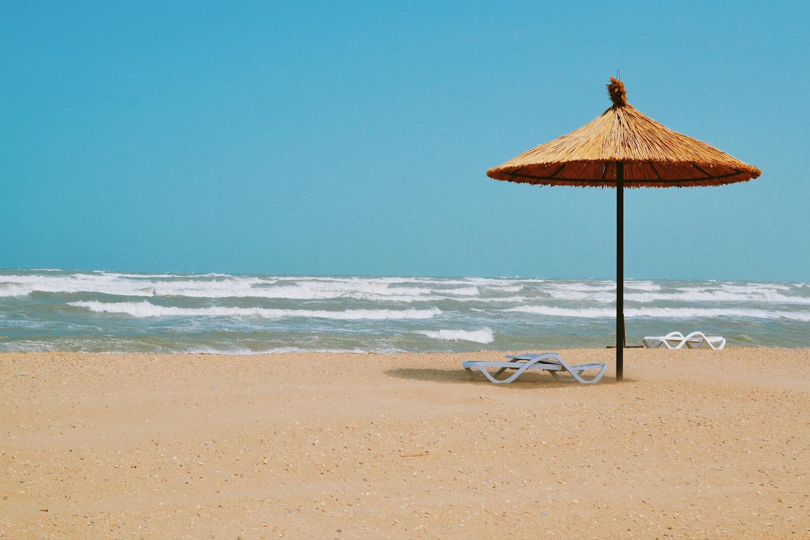 Peaceful sandy beach scene with straw umbrella and empty loungers under a clear blue sky.