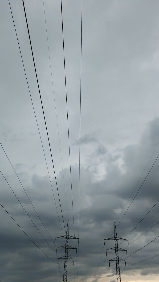 From below power line with poles and wires against gray cloudy sky at dusk