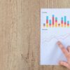 Hand analyzing business graphs on a wooden desk, focusing on data results and growth analysis.