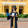 Graduate in cap and gown holding diploma outside historic yellow building.