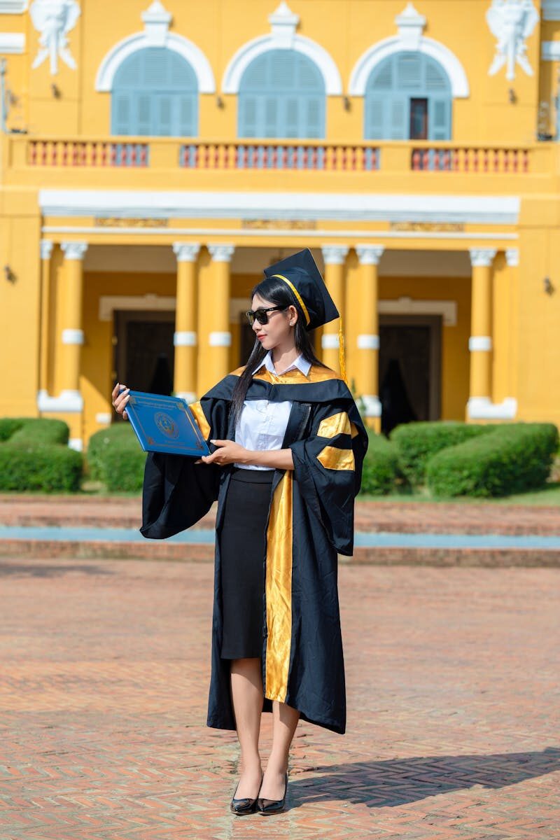Graduate in cap and gown holding diploma outside historic yellow building.