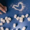 Top view of sugar cubes and a heart drawn in sugar on a blue surface with a hand nearby.