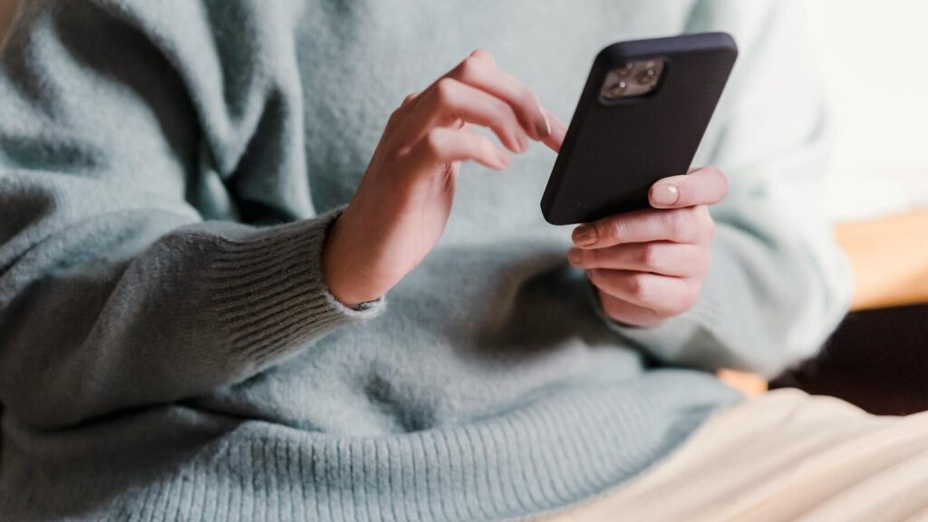 Crop faceless female in casual clothes using smartphone and touching screen while chilling at home