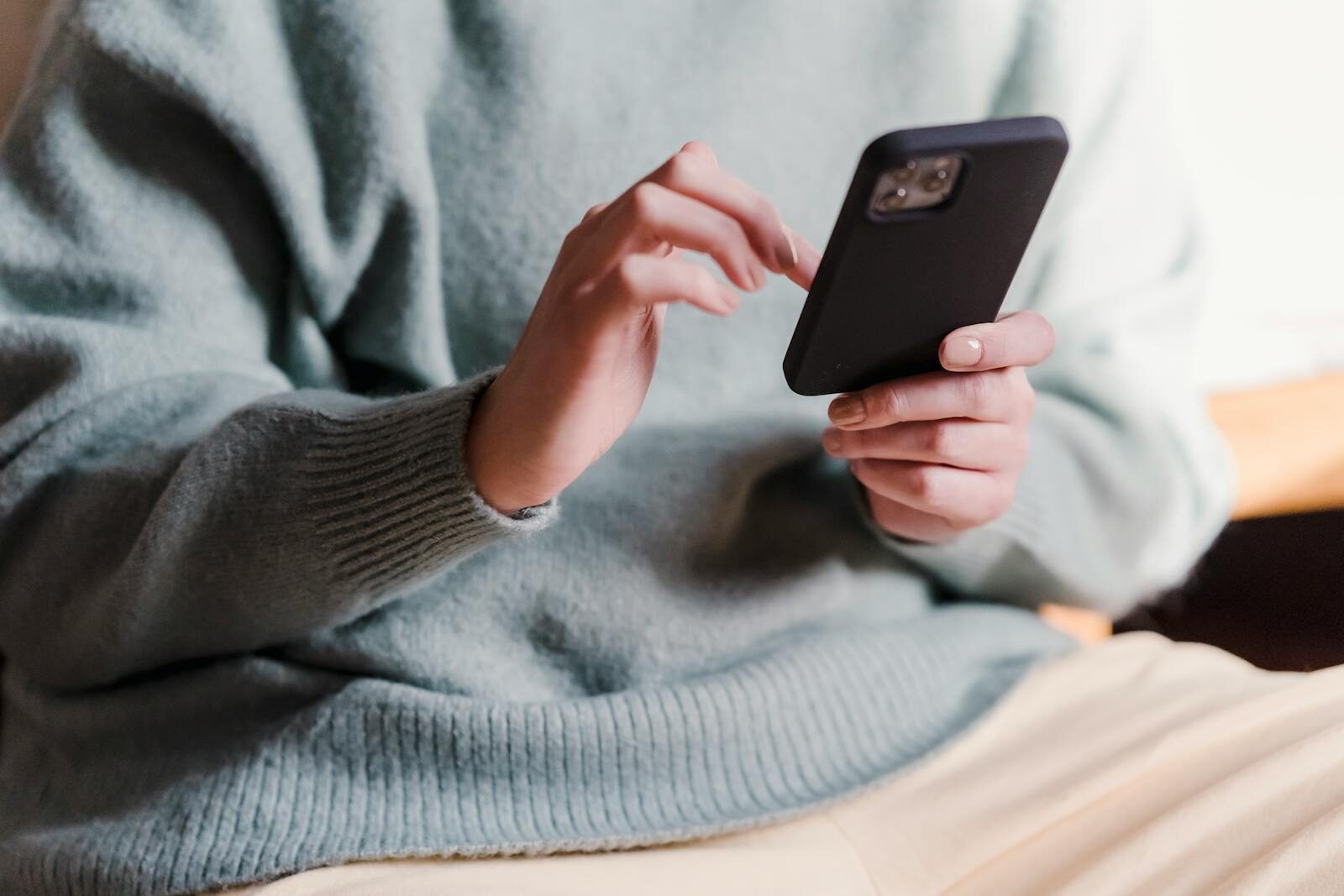 Crop faceless female in casual clothes using smartphone and touching screen while chilling at home