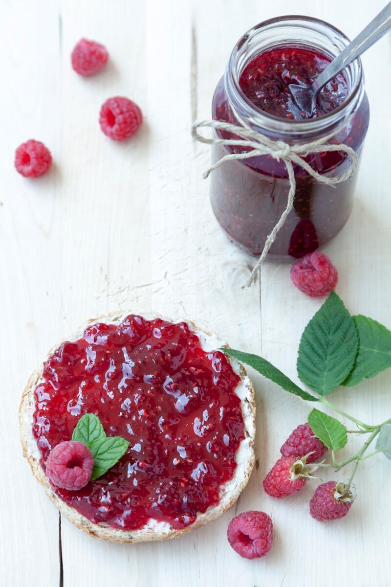 Delicious raspberry jam spread on toast with fresh raspberries on a white wooden background.