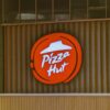 Illuminated Pizza Hut sign on a wooden storefront captured in daylight.