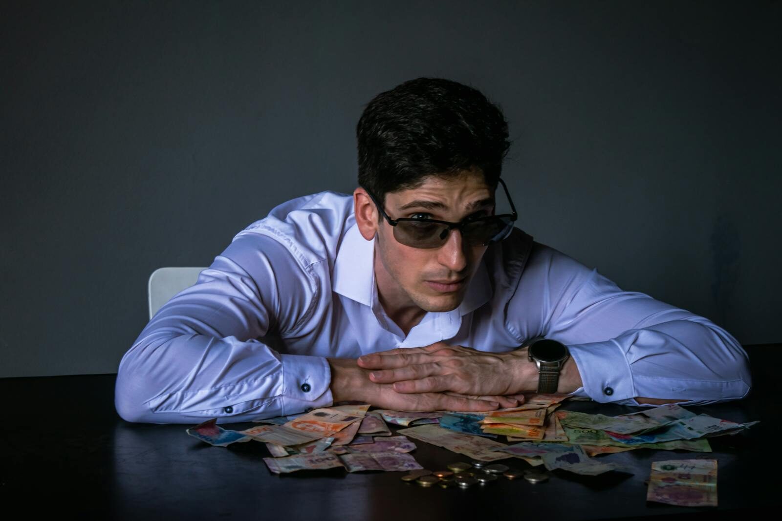 Portrait of a man in sunglasses leaning over a table covered with various currencies.