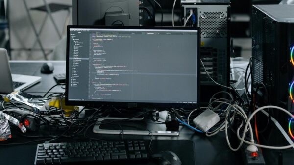 A cluttered workstation in an office featuring a monitor displaying code, surrounded by a keyboard, mouse, and wiring.