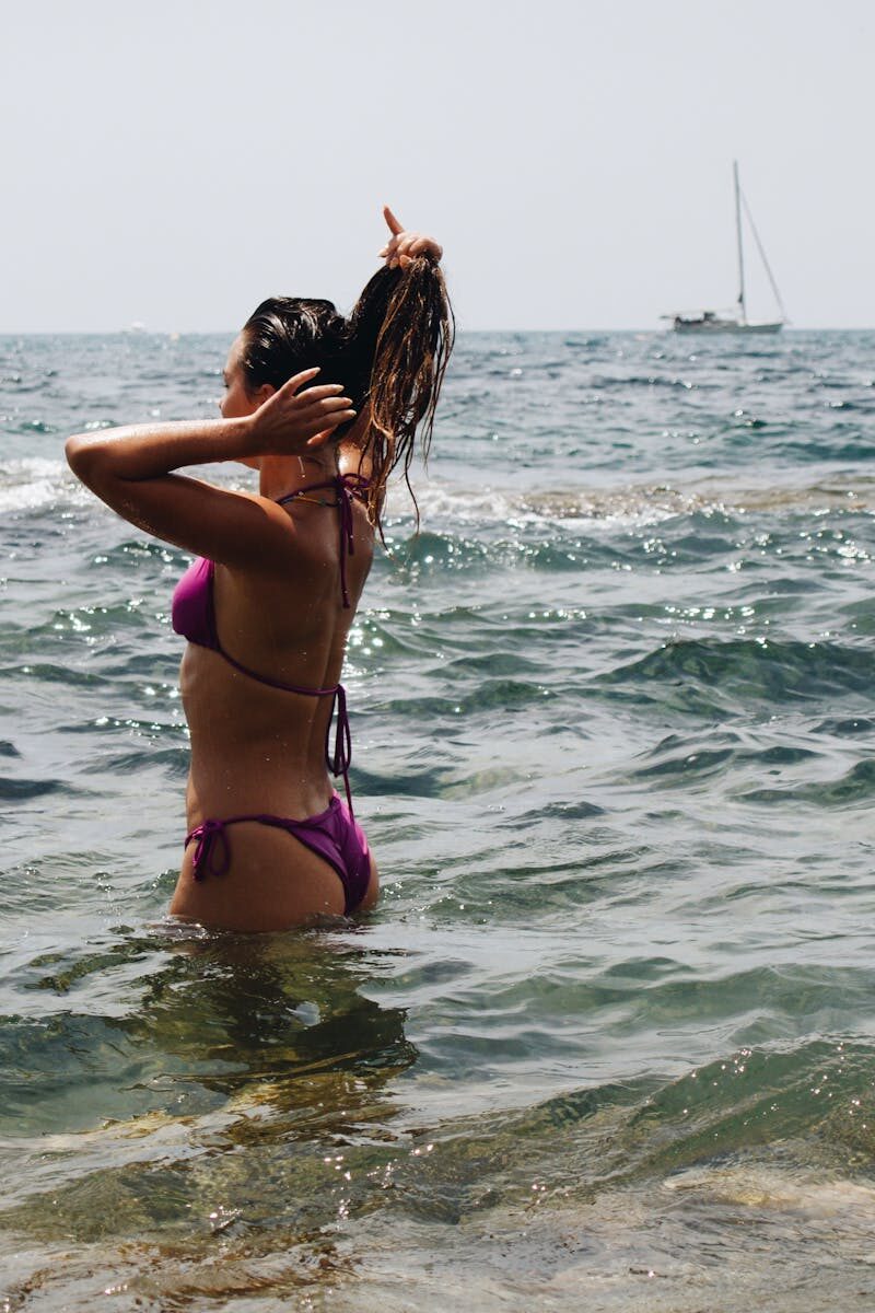 A woman in a bikini enjoying a sunny summer day in the ocean, with a distant sailboat.