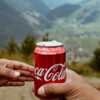 Hands sharing a Coca-Cola can with a scenic mountain backdrop.