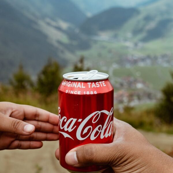 Hands sharing a Coca-Cola can with a scenic mountain backdrop.
