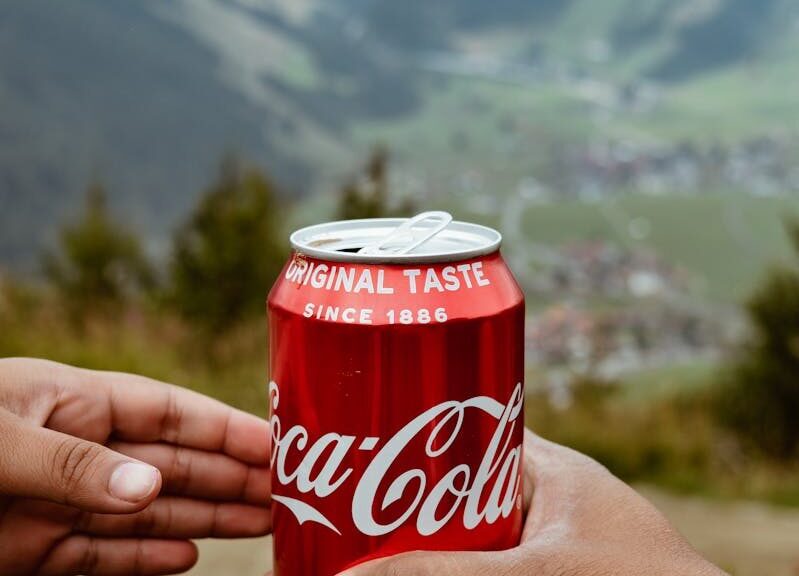 Hands sharing a Coca-Cola can with a scenic mountain backdrop.