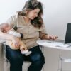 Concentrated mom sitting on chair with cute baby and browsing laptop while working remotely on maternity leave