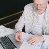 Elderly man with eyeglasses reviewing documents at a laptop. Indoor setting with natural light.