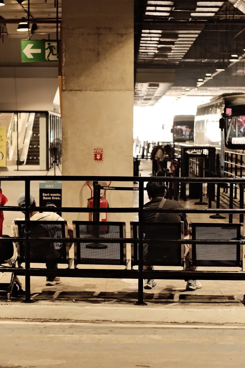 Passengers waiting at a bus station with visible signs and buses in the background.