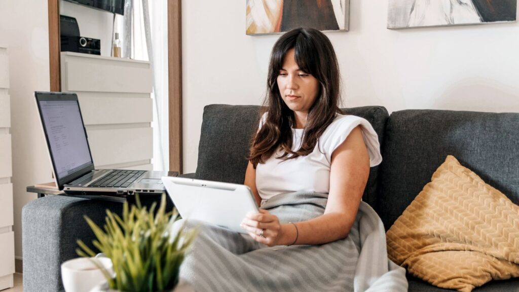 Woman on sofa with tablet and laptop, cozy home work environment.