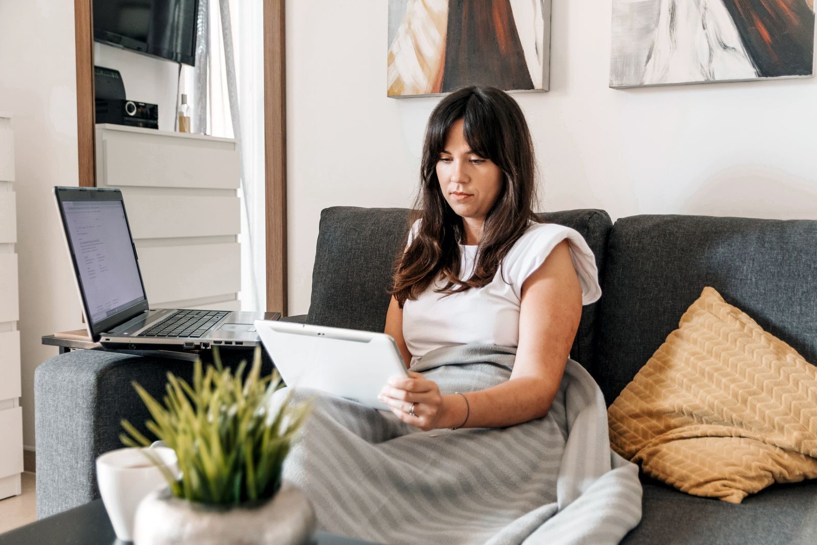 Woman on sofa with tablet and laptop, cozy home work environment.