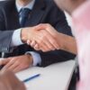 Two businessmen shaking hands across table, symbolizing agreement and partnership in an office environment.