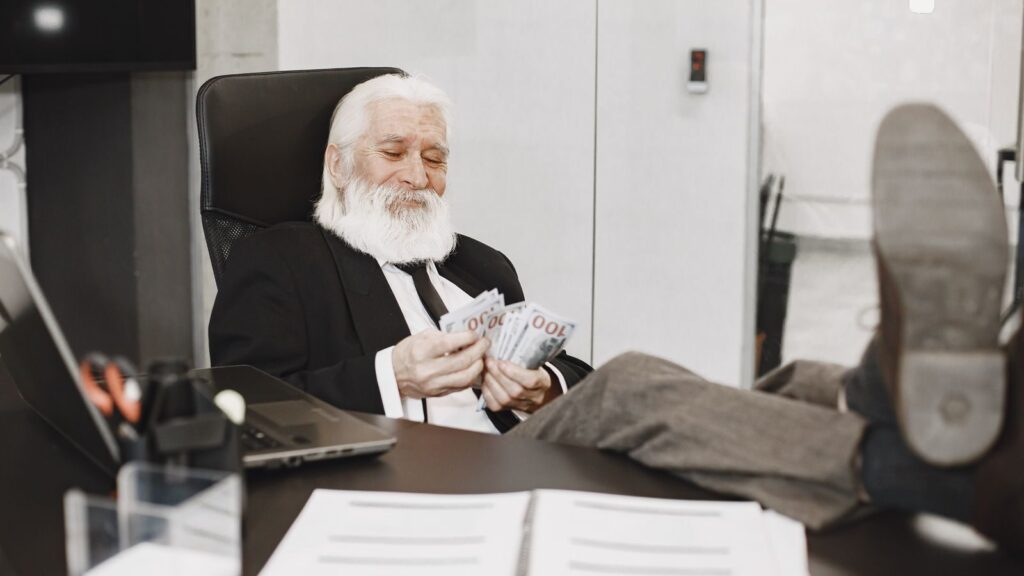 Elderly businessman relaxing at desk, counting money notes in a modern office setting.