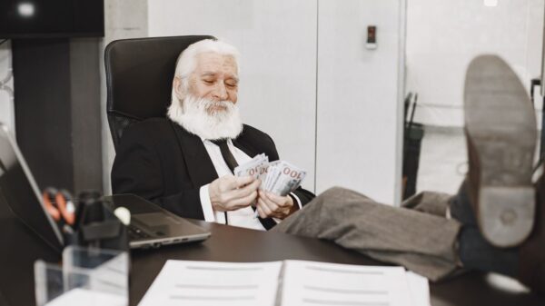 Elderly businessman relaxing at desk, counting money notes in a modern office setting.