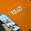 Hand holding smartphone with Temu app beside orange Temu package.