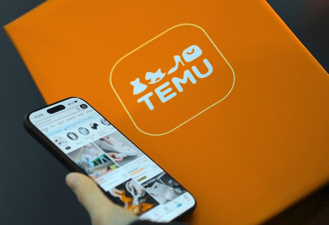 Hand holding smartphone with Temu app beside orange Temu package.