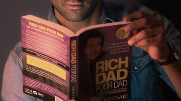 Portrait of a young man intently reading 'Rich Dad Poor Dad' against a gray wall.