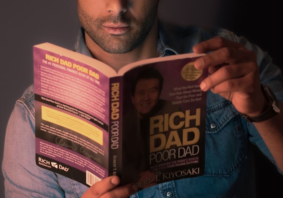 Portrait of a young man intently reading 'Rich Dad Poor Dad' against a gray wall.