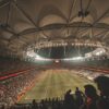 From above of spectators sitting in stands while observing empty green rectangular soccer field under artificial lights before match