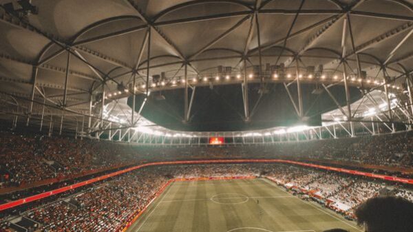 From above of spectators sitting in stands while observing empty green rectangular soccer field under artificial lights before match