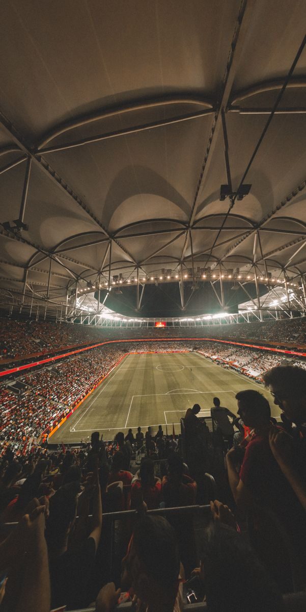 From above of spectators sitting in stands while observing empty green rectangular soccer field under artificial lights before match