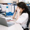 A woman in an office sitting at a desk, visibly stressed, using a phone. Workplace stress concept.