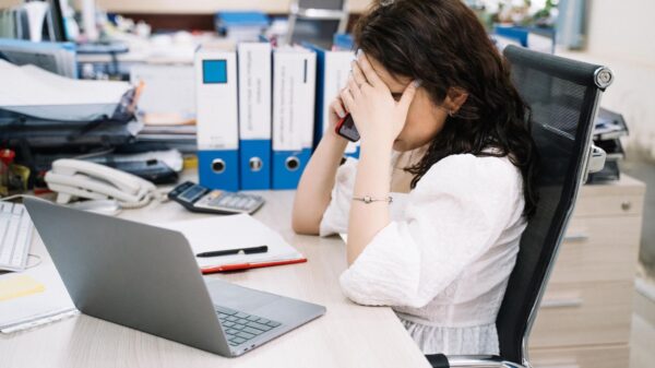 A woman in an office sitting at a desk, visibly stressed, using a phone. Workplace stress concept.