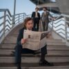 Young professionals in suits, one reading a newspaper on stairs outdoors.