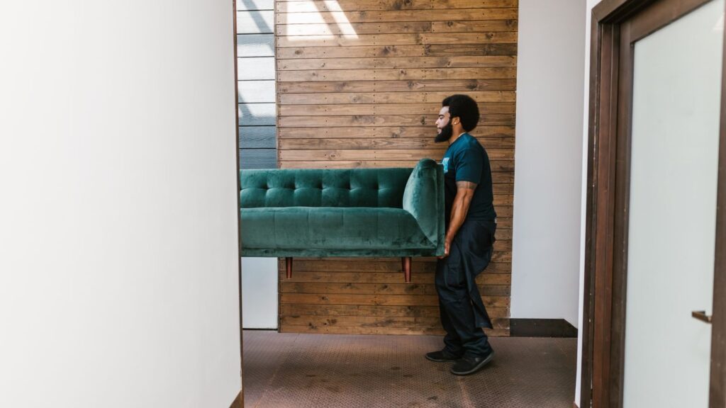 A mover carries a green sofa indoors, showcasing modern interior design and relocation services.