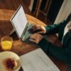 Woman typing on laptop at wooden table with breakfast.