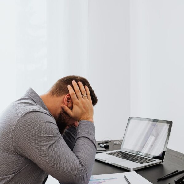 A frustrated man sits at a desk with a laptop and charts, showing work stress.