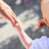 Blonde child holding parent's hand outdoors, symbolizing love and protection.