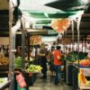 A bustling outdoor farmer's market with fresh vegetables and people shopping on a sunny day.