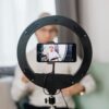 Seated man vlogging with a smartphone on a ring light tripod indoors.