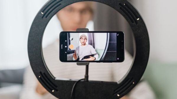 Seated man vlogging with a smartphone on a ring light tripod indoors.