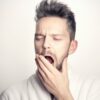 Close-up of a bearded man yawning with hand on mouth against a white background.