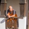 An elderly man in work overalls leaning on a vintage wooden railing.