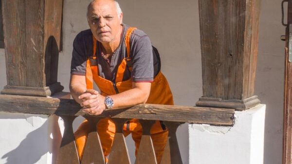 An elderly man in work overalls leaning on a vintage wooden railing.