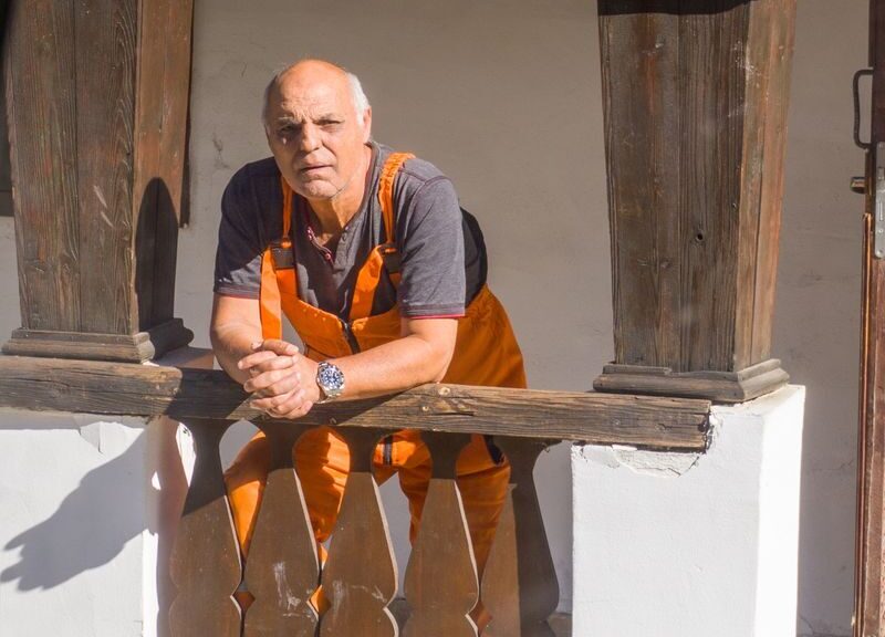 An elderly man in work overalls leaning on a vintage wooden railing.